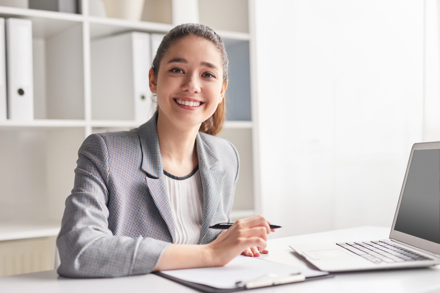 Happy young female smiling and looking at camera while sitting at desk and making notes in clipboard in office