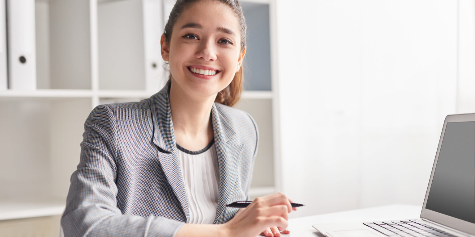 Happy young female smiling and looking at camera while sitting at desk and making notes in clipboard in office